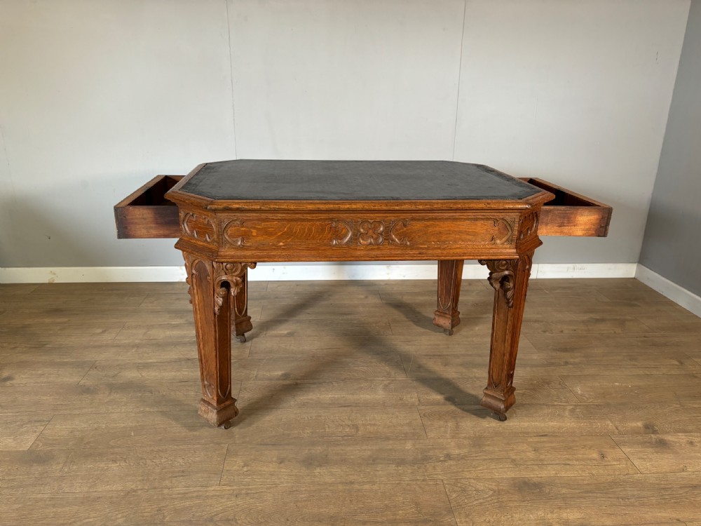 A Victorian Gothic Revival oak writing table featuring a dark leather inset top, carved quatrefoil frieze details, and detailed corner legs on castors. View X