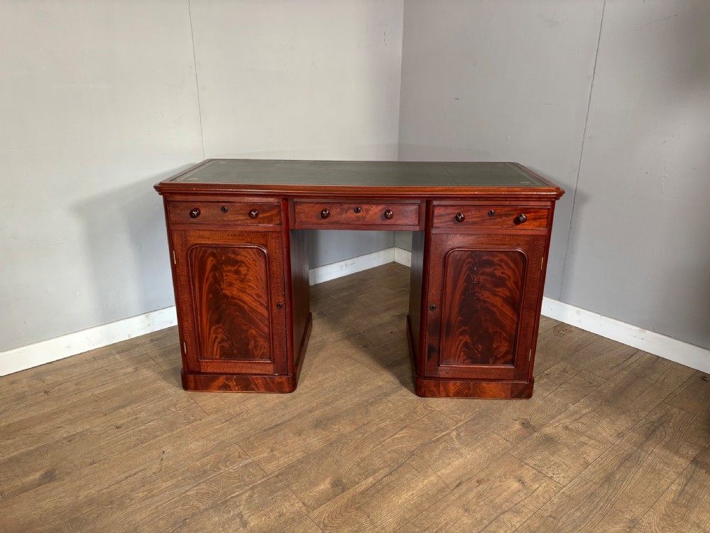 Victorian mahogany kneehole desk featuring a dark green leather writing surface, central frieze drawer, turned wooden knobs, and two cupboard pedestals with flame mahogany veneer panels.
