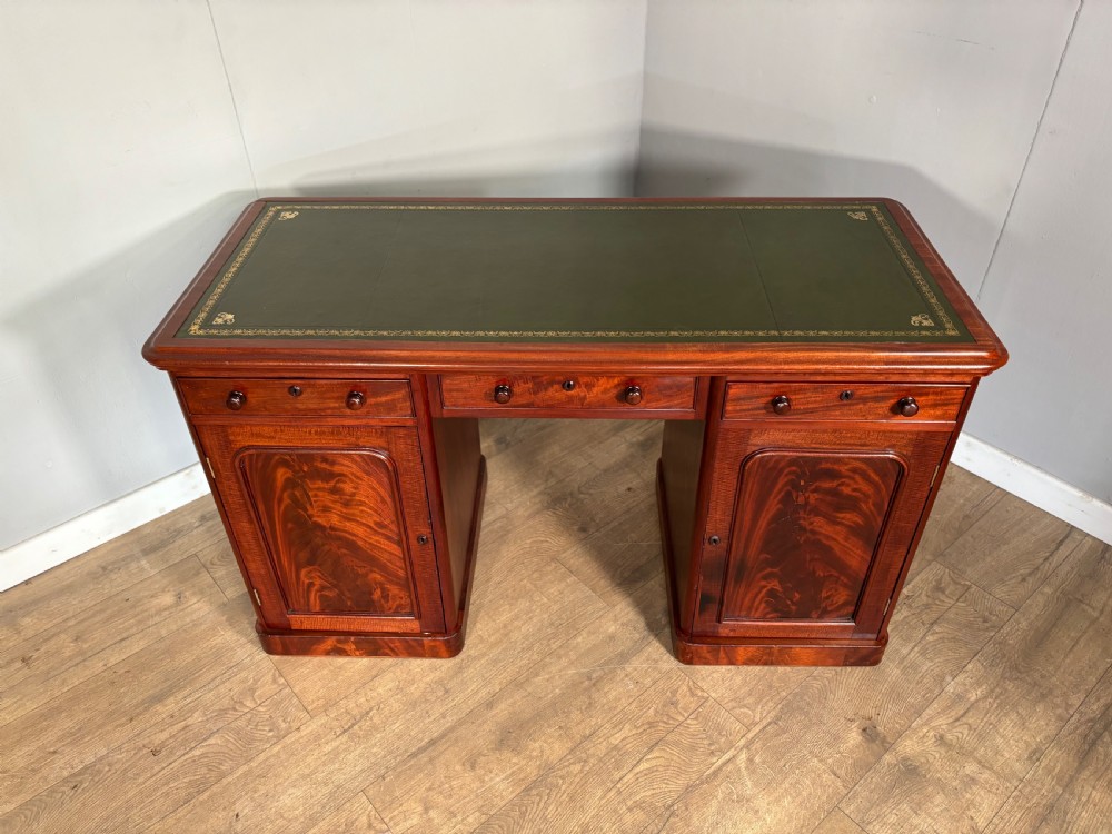 Victorian mahogany kneehole desk featuring a dark green leather writing surface, central frieze drawer, turned wooden knobs, and two cupboard pedestals with flame mahogany veneer panels. View II