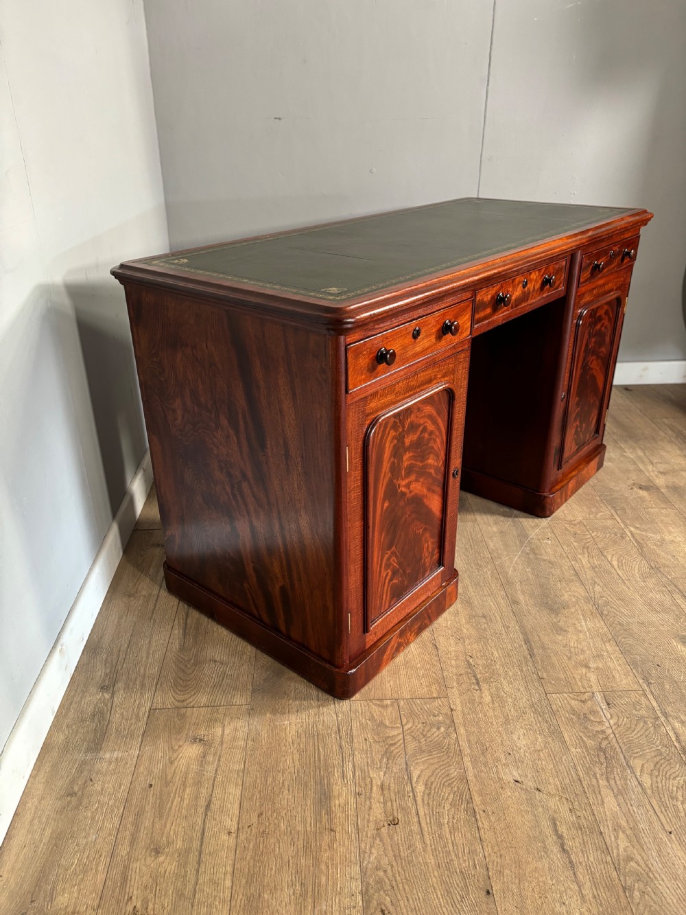 Victorian mahogany kneehole desk featuring a dark green leather writing surface, central frieze drawer, turned wooden knobs, and two cupboard pedestals with flame mahogany veneer panels. View III