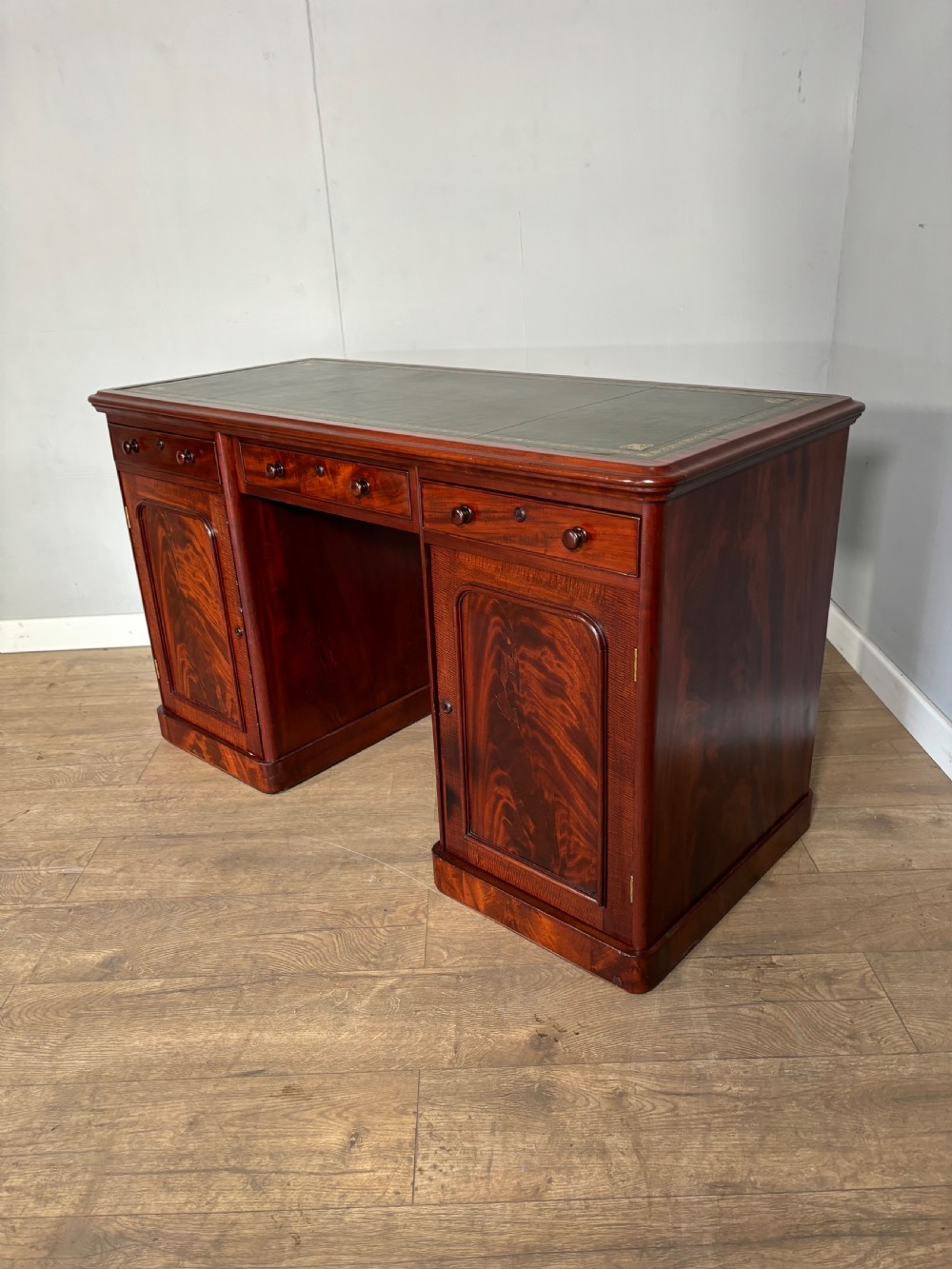 Victorian mahogany kneehole desk featuring a dark green leather writing surface, central frieze drawer, turned wooden knobs, and two cupboard pedestals with flame mahogany veneer panels. View V