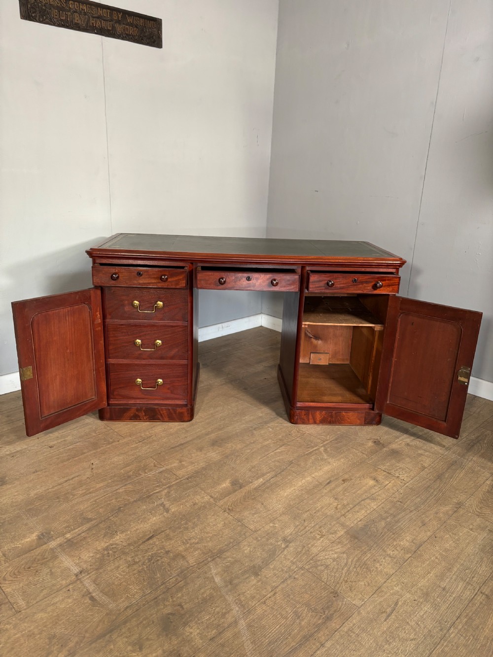 Victorian mahogany kneehole desk featuring a dark green leather writing surface, central frieze drawer, turned wooden knobs, and two cupboard pedestals with flame mahogany veneer panels. View VI