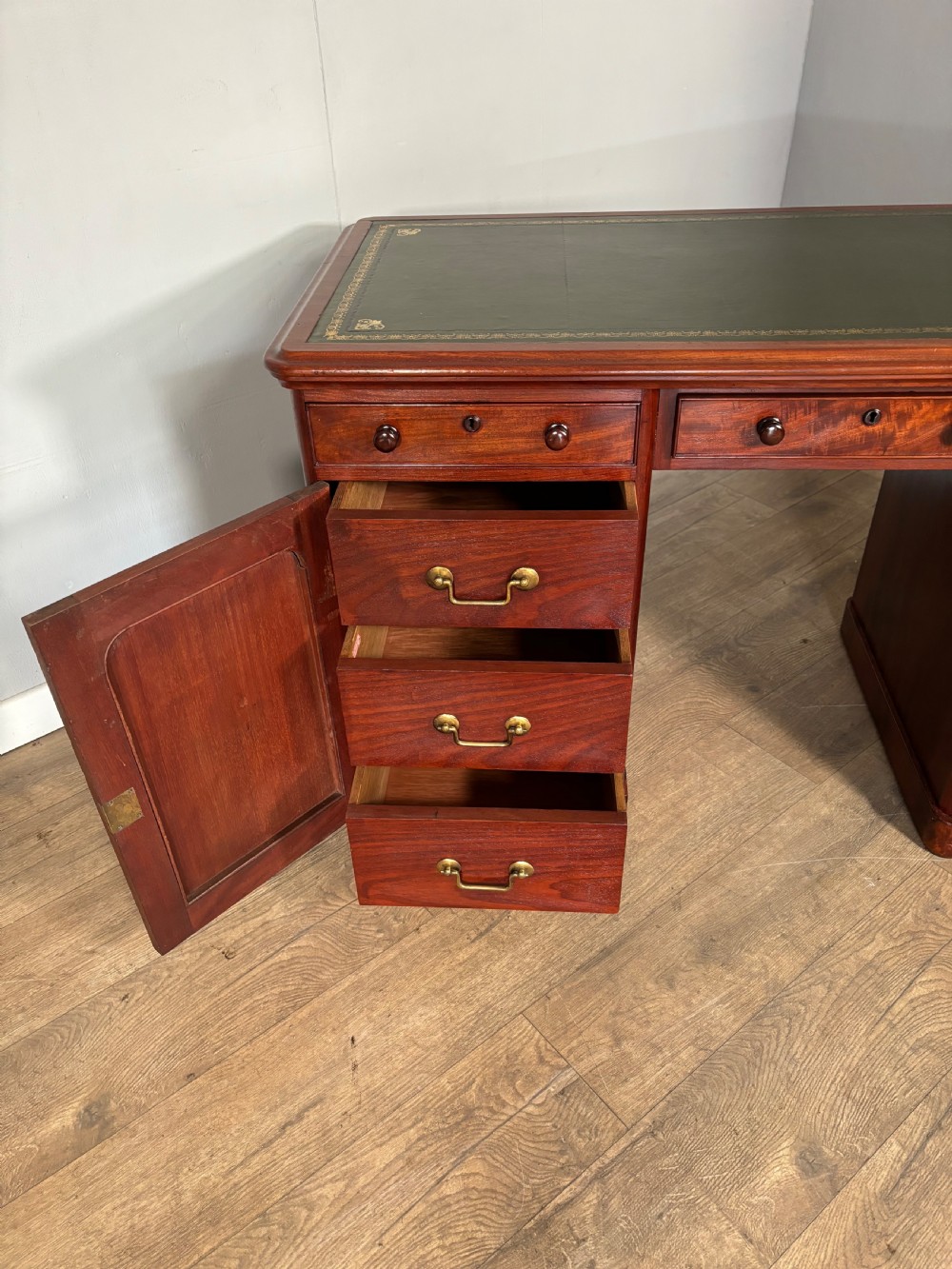 Victorian mahogany kneehole desk featuring a dark green leather writing surface, central frieze drawer, turned wooden knobs, and two cupboard pedestals with flame mahogany veneer panels. View VII