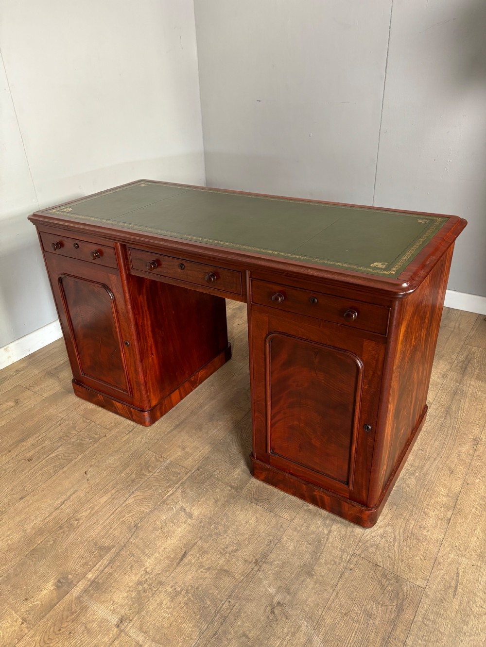 Victorian mahogany kneehole desk featuring a dark green leather writing surface, central frieze drawer, turned wooden knobs, and two cupboard pedestals with flame mahogany veneer panels. View IX