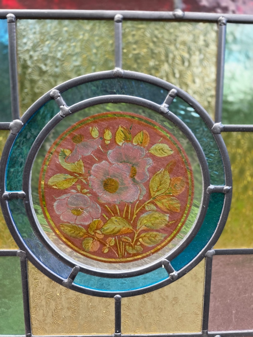 A rectangular leaded glass panel framed in light pine featuring a central hand-painted pink floral motif surrounded by green, red, amber bullseye, and textured glass sections. View IV
