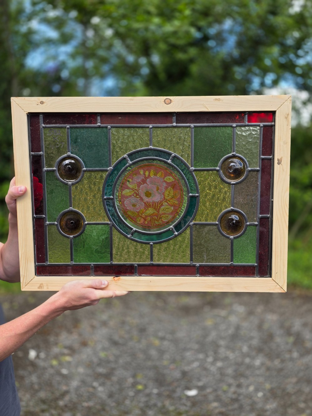 A rectangular leaded glass panel framed in light pine featuring a central hand-painted pink floral motif surrounded by green, red, amber bullseye, and textured glass sections. View II