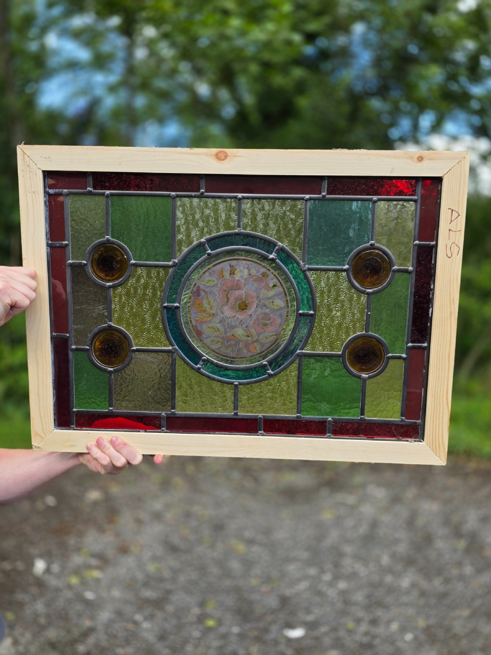A rectangular leaded glass panel framed in light pine featuring a central hand-painted pink floral motif surrounded by green, red, amber bullseye, and textured glass sections. View III