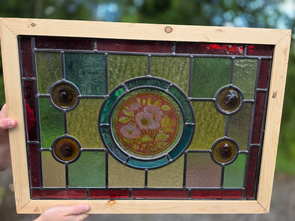 A rectangular leaded glass panel framed in light pine featuring a central hand-painted pink floral motif surrounded by green, red, amber bullseye, and textured glass sections.