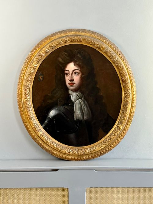 fig 1. An oval late 17th-century oil portrait of a long-haired nobleman in dark plate armour with a white cravat, set within a carved gilt frame.