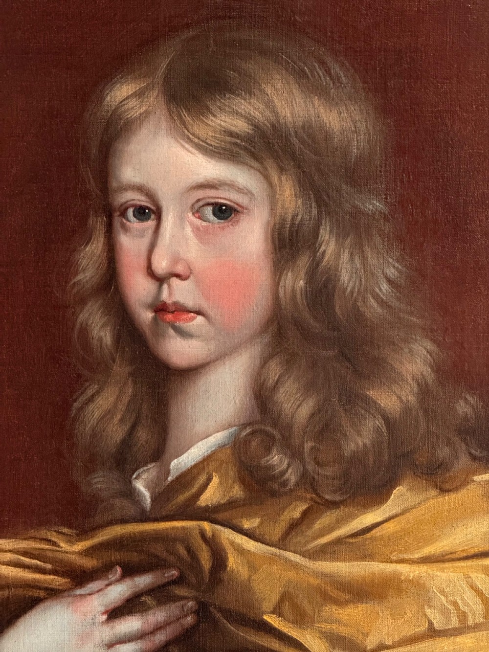 A 17th-century oil portrait of a young boy with long curled hair draped in golden-yellow silk, set within a painted trompe loeil oval border. View III