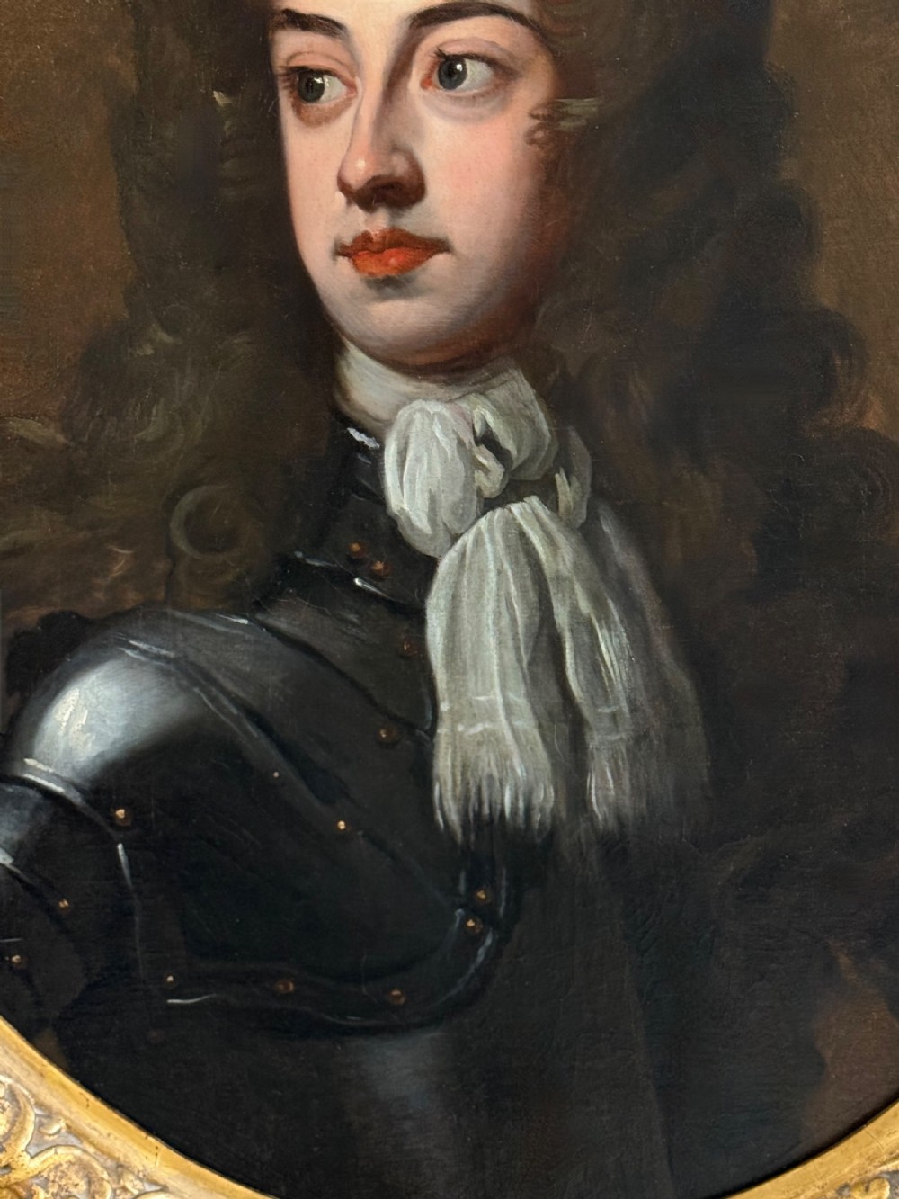 An oval late 17th-century oil portrait of a long-haired nobleman in dark plate armour with a white cravat, set within a carved gilt frame. View VII