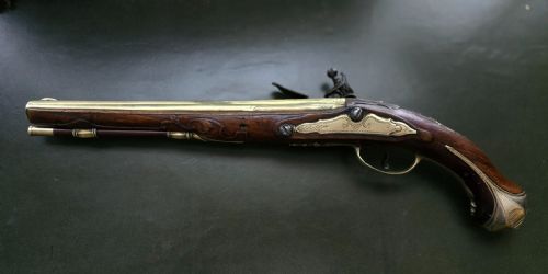 fig 1. An 18th-century French flintlock holster pistol featuring a carved walnut stock, polished brass barrel, engraved brass lockplate, and steel mechanism.