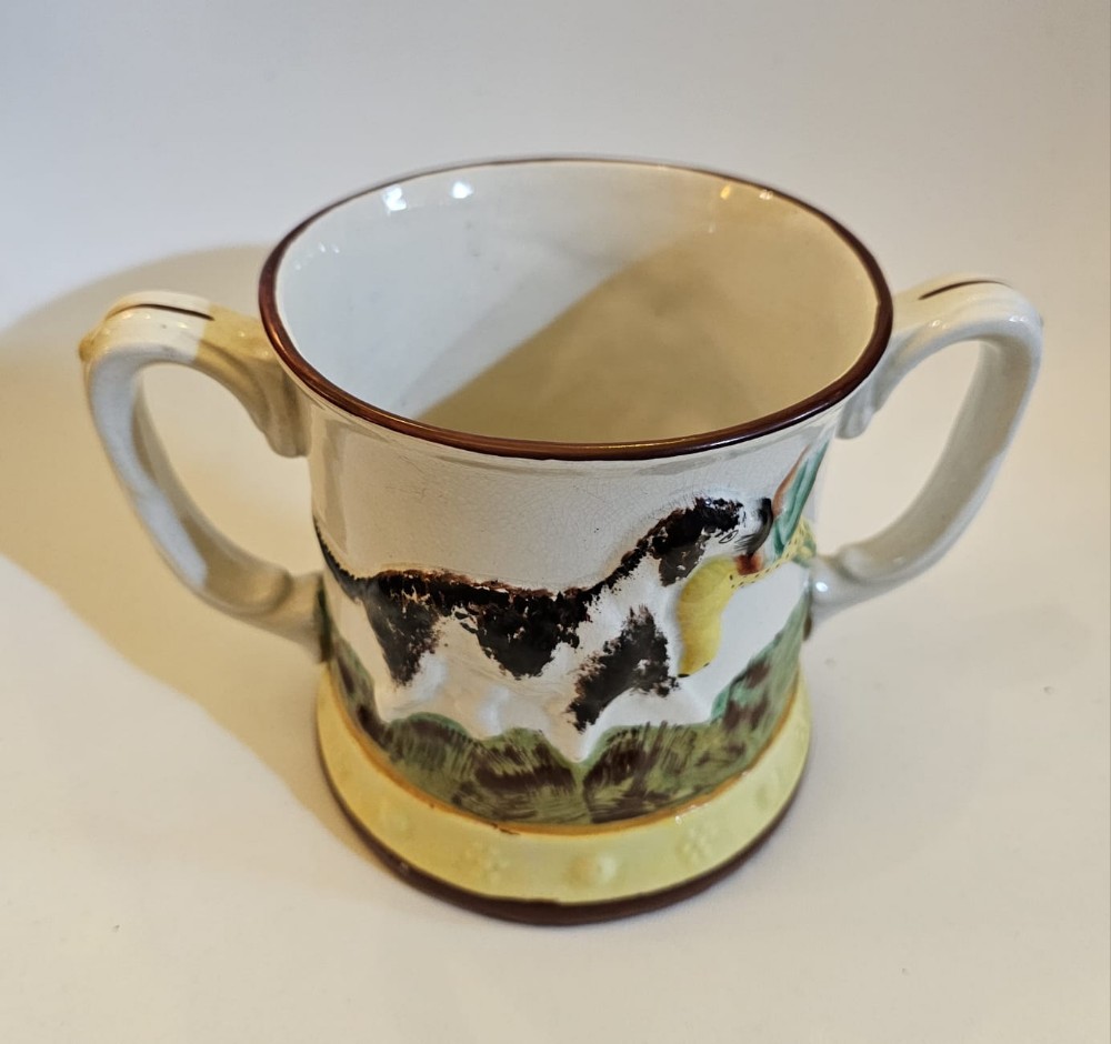 An antique Staffordshire pottery frog mug featuring a painted hunting dog motif against a pale background with two scrolled handles.