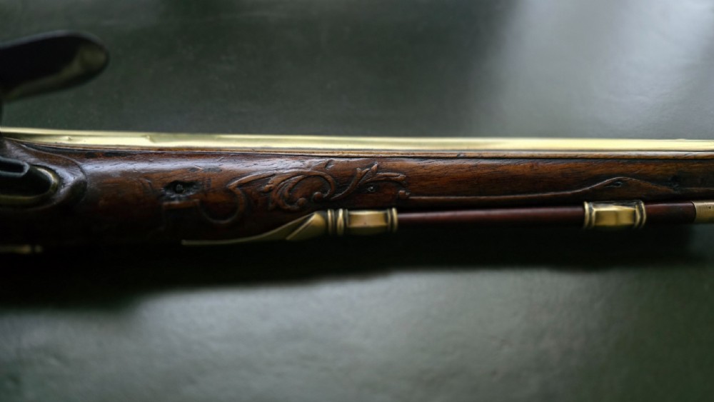 An 18th-century French flintlock holster pistol featuring a carved walnut stock, polished brass barrel, engraved brass lockplate, and steel mechanism. View IV