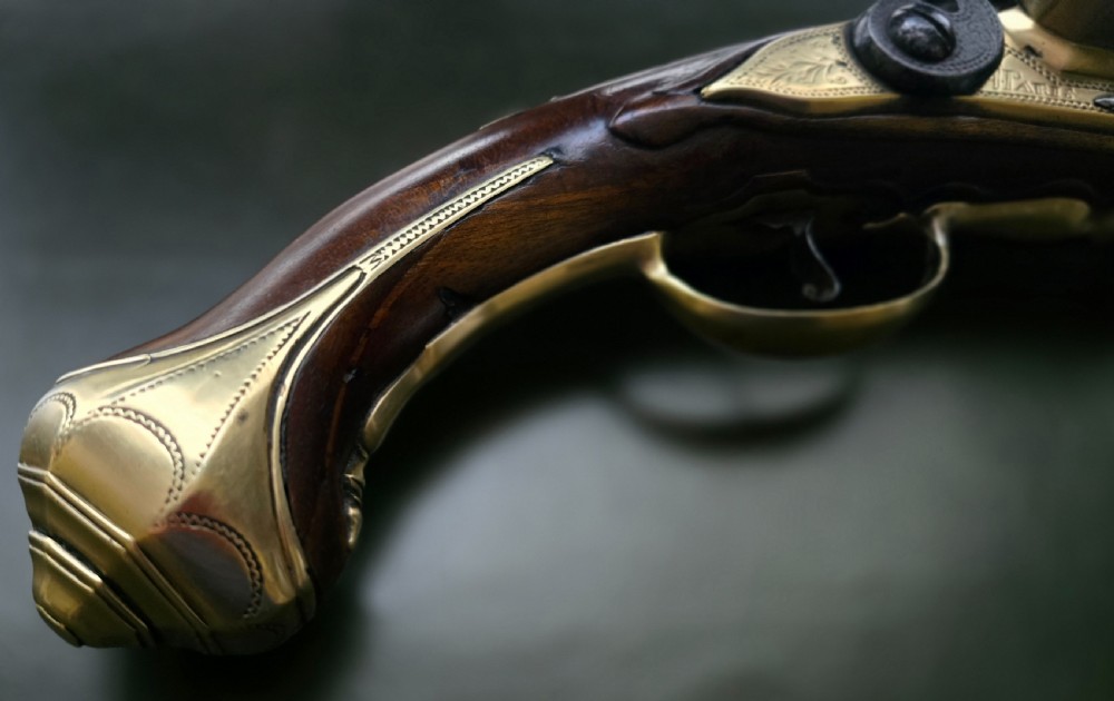 An 18th-century French flintlock holster pistol featuring a carved walnut stock, polished brass barrel, engraved brass lockplate, and steel mechanism. View V
