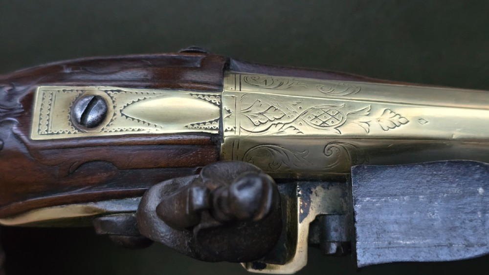 An 18th-century French flintlock holster pistol featuring a carved walnut stock, polished brass barrel, engraved brass lockplate, and steel mechanism. View X