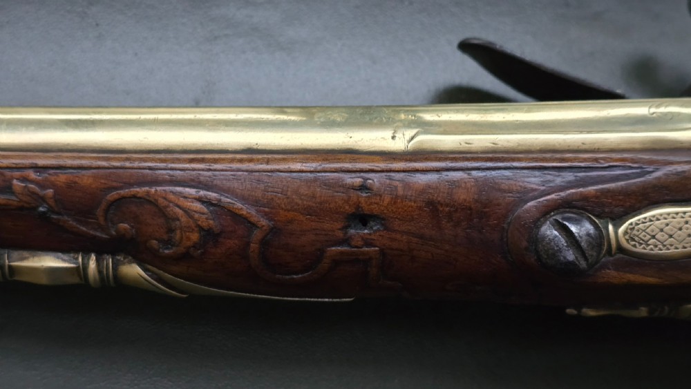 An 18th-century French flintlock holster pistol featuring a carved walnut stock, polished brass barrel, engraved brass lockplate, and steel mechanism. View XX