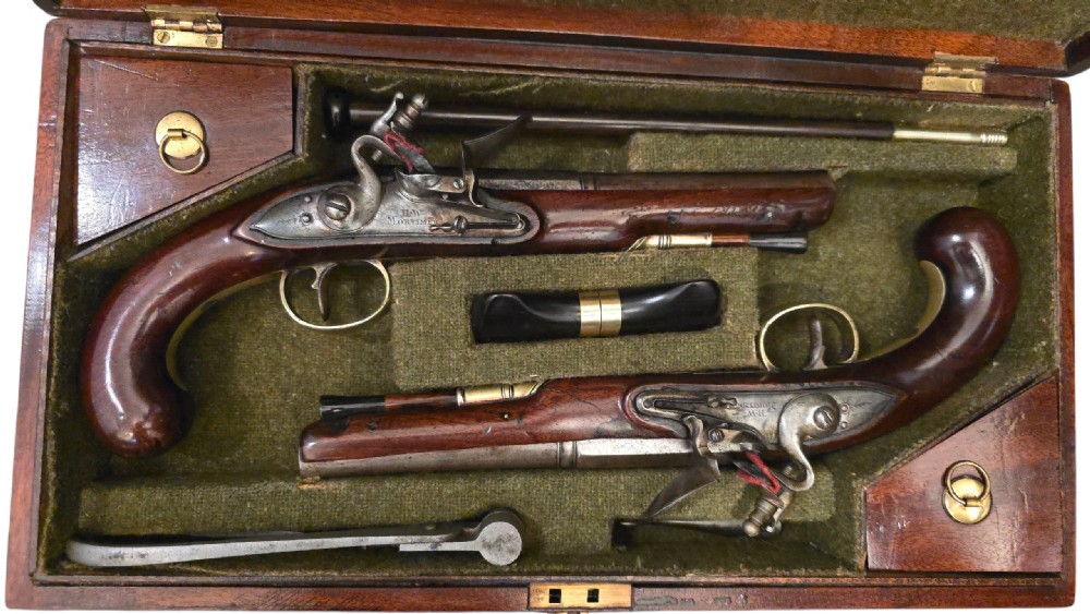A fitted mahogany wooden case lined with green baize containing a pair of flintlock duelling pistols, a ramrod, and loading accessories.