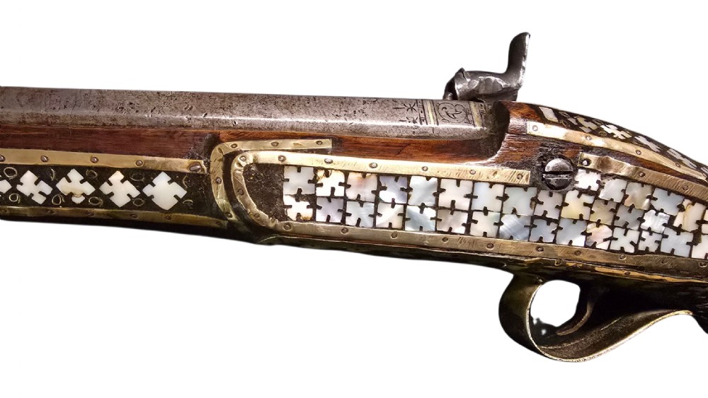 A 19th-century Ottoman pistol featuring a dark wood stock inlaid with mother-of-pearl geometric designs and brass wire, with an engraved steel mechanism and barrel. View V