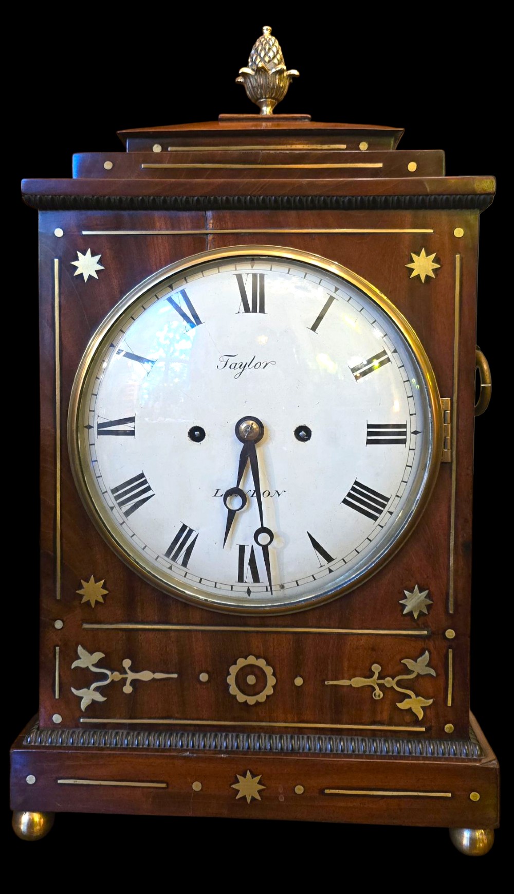 A Georgian mahogany bracket clock featuring brass star and floral motifs, a pineapple finial, brass ball feet, and a white dial marked Taylor London with Roman numerals. View XII