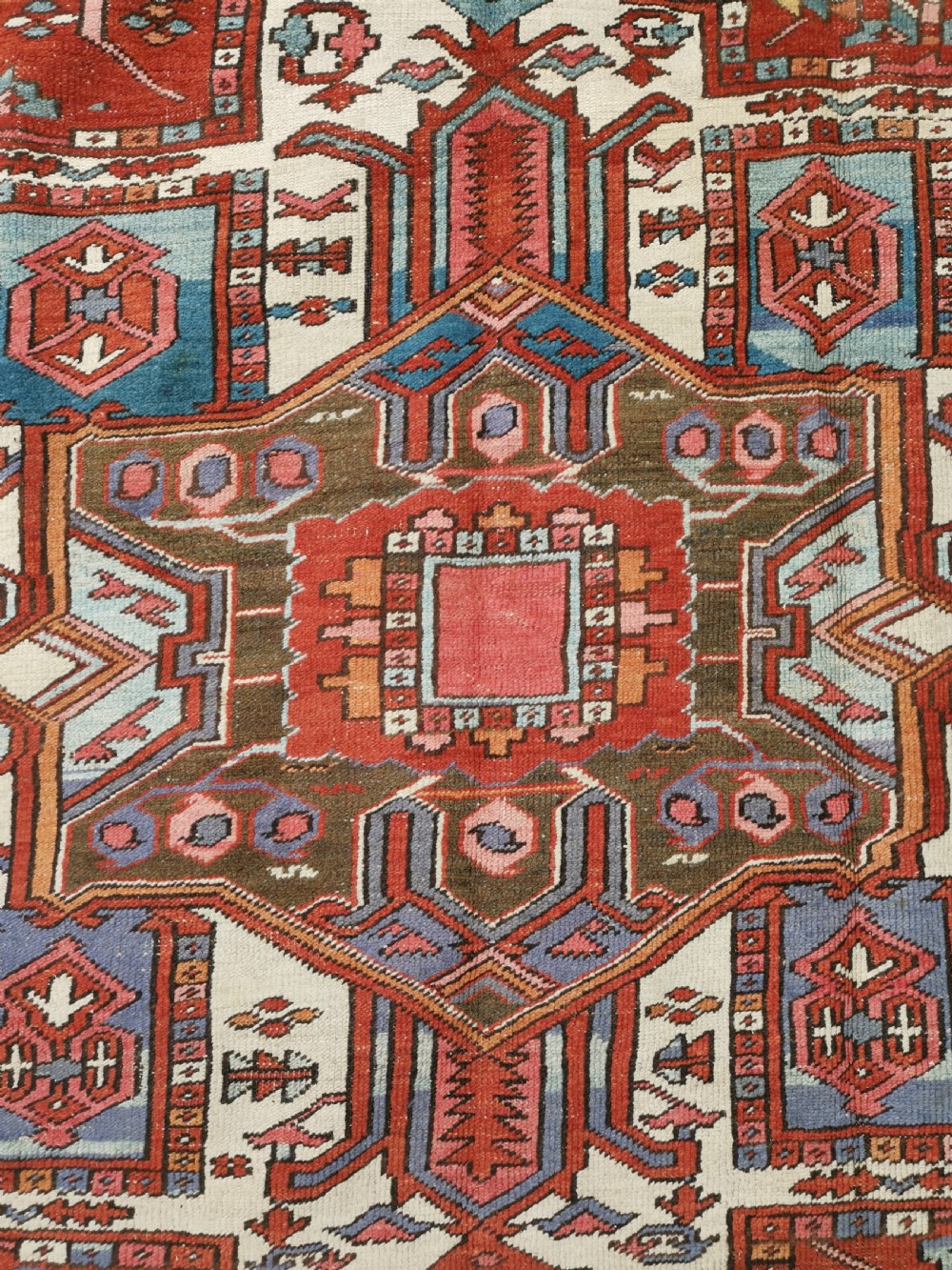 A rectangular Persian Heriz hand-knotted wool carpet featuring a red geometric field with a central star medallion, cream corner spandrels, and multi-banded borders. View V