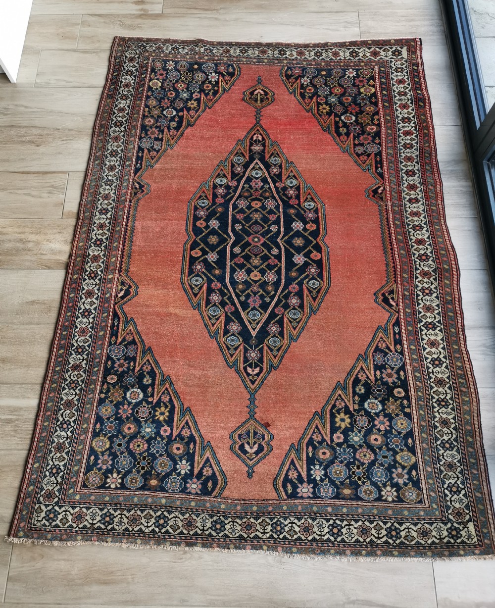 An antique Persian Malayer wool rug featuring a terracotta field, a central navy blue diamond medallion, and detailed geometric floral borders.