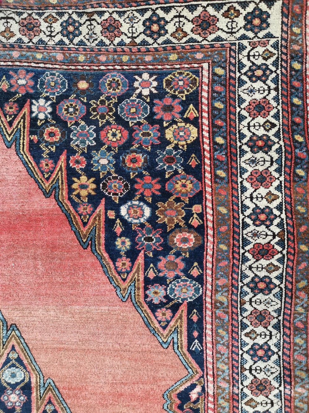 An antique Persian Malayer wool rug featuring a terracotta field, a central navy blue diamond medallion, and detailed geometric floral borders. View IV