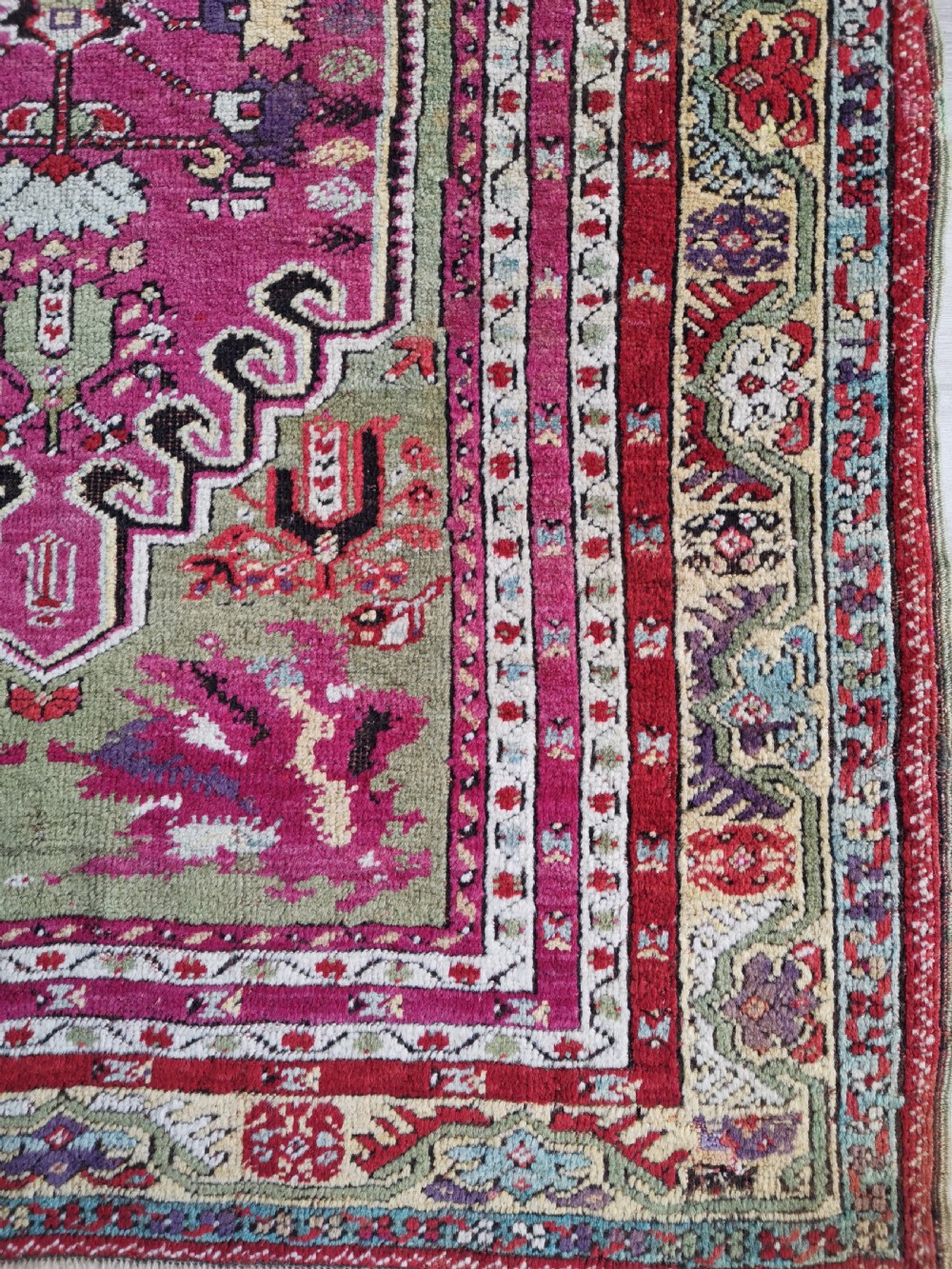 A hand-knotted Anatolian Kirsehir wool rug featuring a central magenta stepped medallion set within an olive green field and surrounded by multi-banded geometric borders. View V