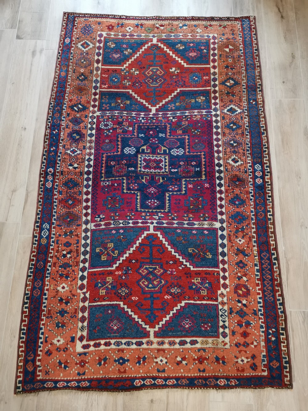 A rectangular hand-knotted wool rug featuring geometric stepped medallions and stylized tribal motifs in shades of red, indigo blue, terracotta, and off-white.