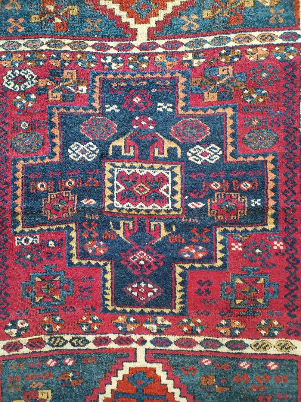 A rectangular hand-knotted wool rug featuring geometric stepped medallions and stylized tribal motifs in shades of red, indigo blue, terracotta, and off-white. View II