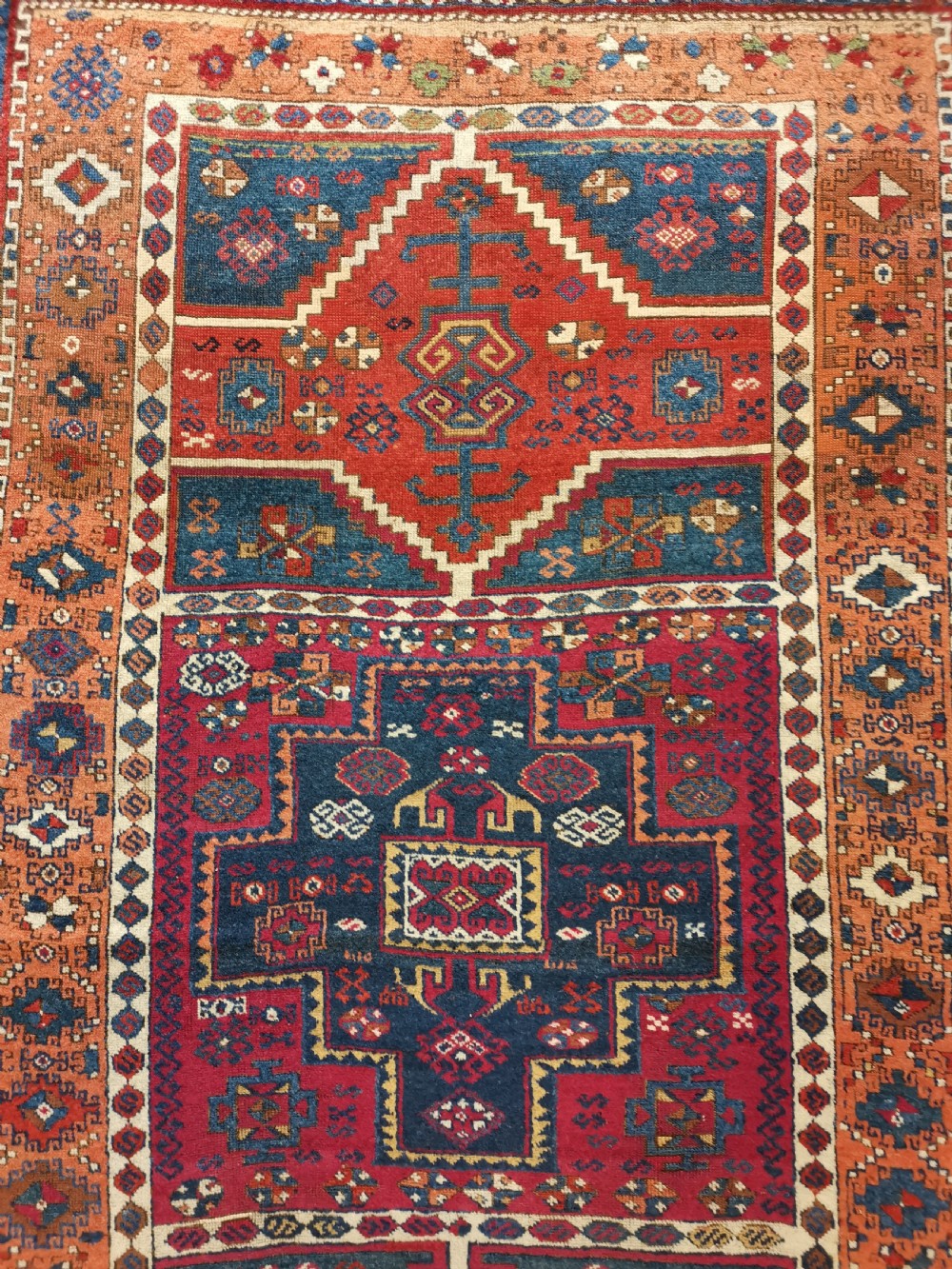 A rectangular hand-knotted wool rug featuring geometric stepped medallions and stylized tribal motifs in shades of red, indigo blue, terracotta, and off-white. View III