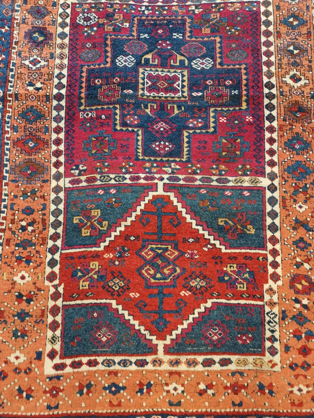 A rectangular hand-knotted wool rug featuring geometric stepped medallions and stylized tribal motifs in shades of red, indigo blue, terracotta, and off-white. View IV