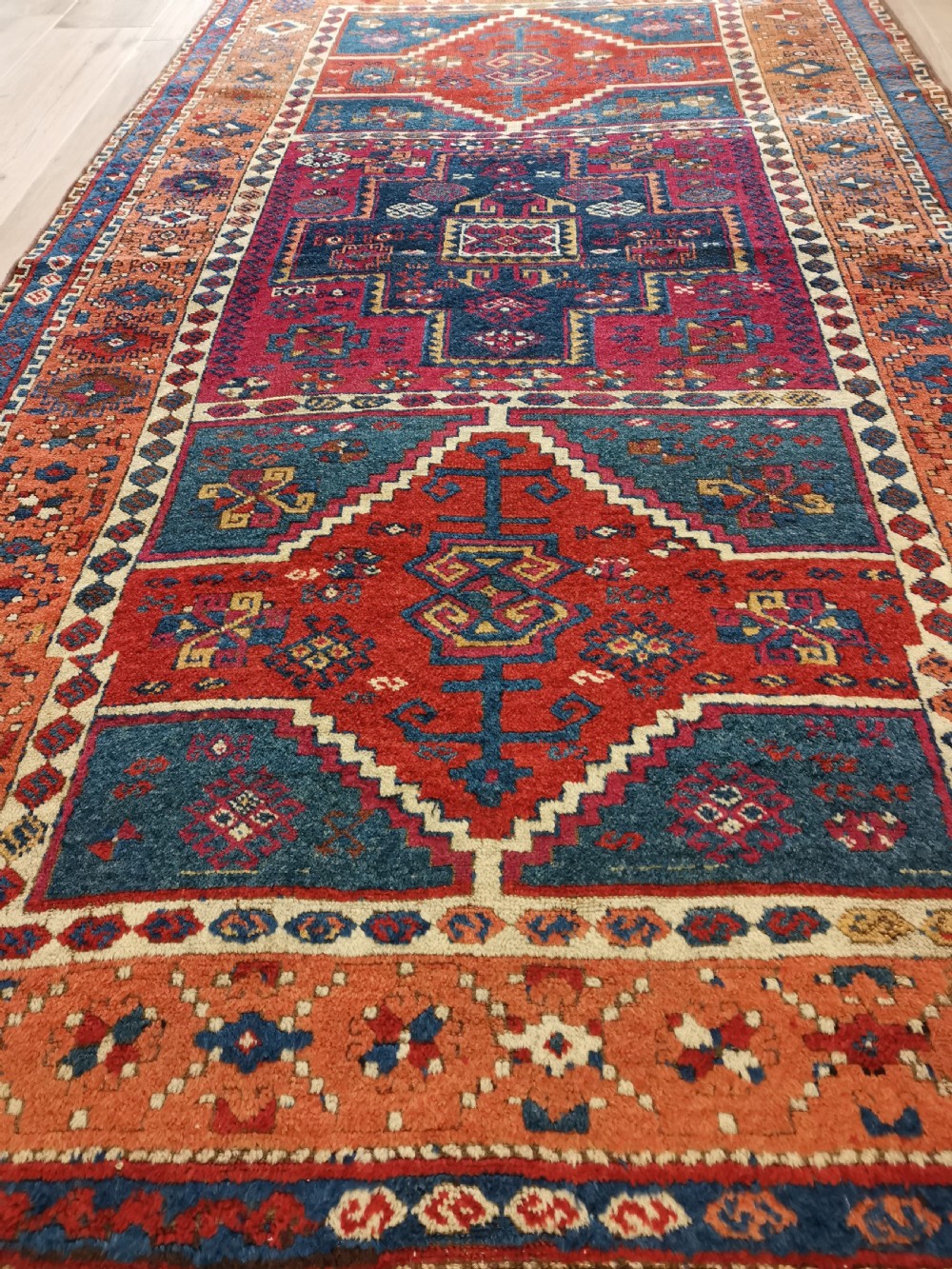 A rectangular hand-knotted wool rug featuring geometric stepped medallions and stylized tribal motifs in shades of red, indigo blue, terracotta, and off-white. View V