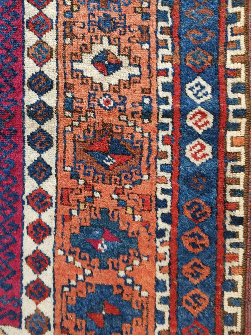 A rectangular hand-knotted wool rug featuring geometric stepped medallions and stylized tribal motifs in shades of red, indigo blue, terracotta, and off-white. View VI