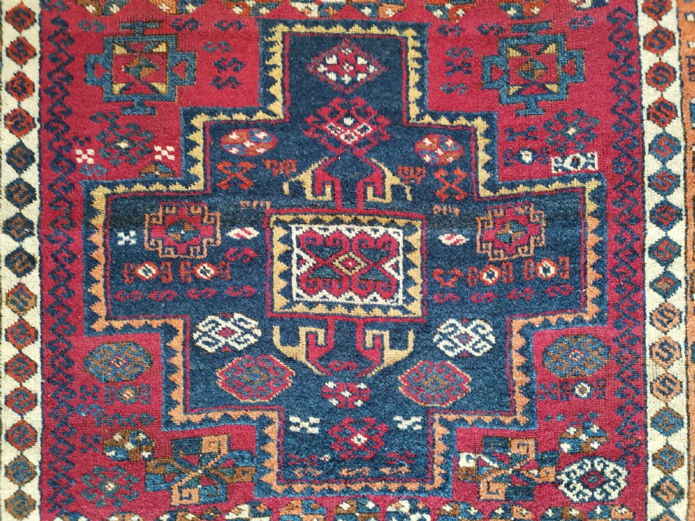 A rectangular hand-knotted wool rug featuring geometric stepped medallions and stylized tribal motifs in shades of red, indigo blue, terracotta, and off-white. View VII