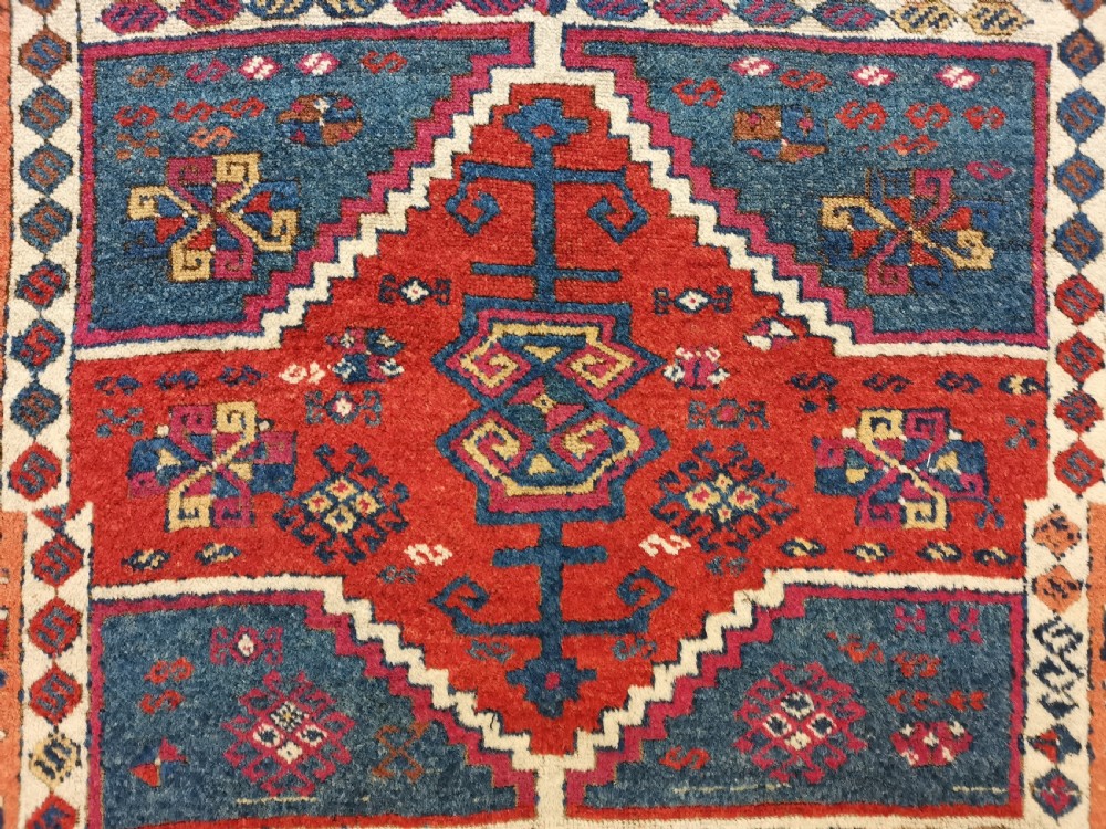 A rectangular hand-knotted wool rug featuring geometric stepped medallions and stylized tribal motifs in shades of red, indigo blue, terracotta, and off-white. View IX