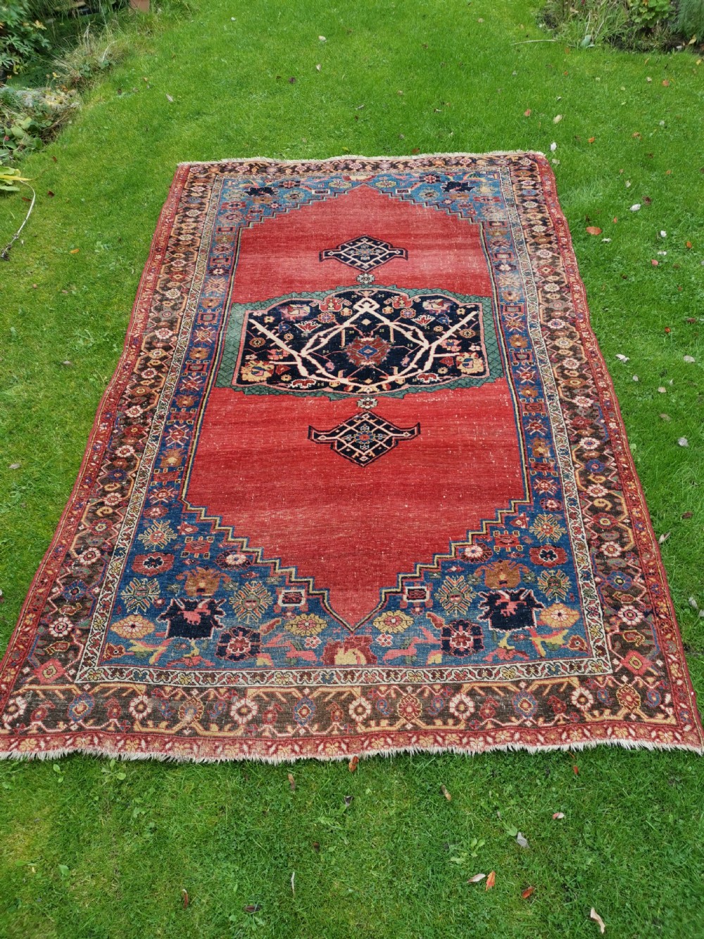 An antique hand-knotted Persian Bijar wool rug featuring a red central field, dark navy medallion, and geometric floral borders.