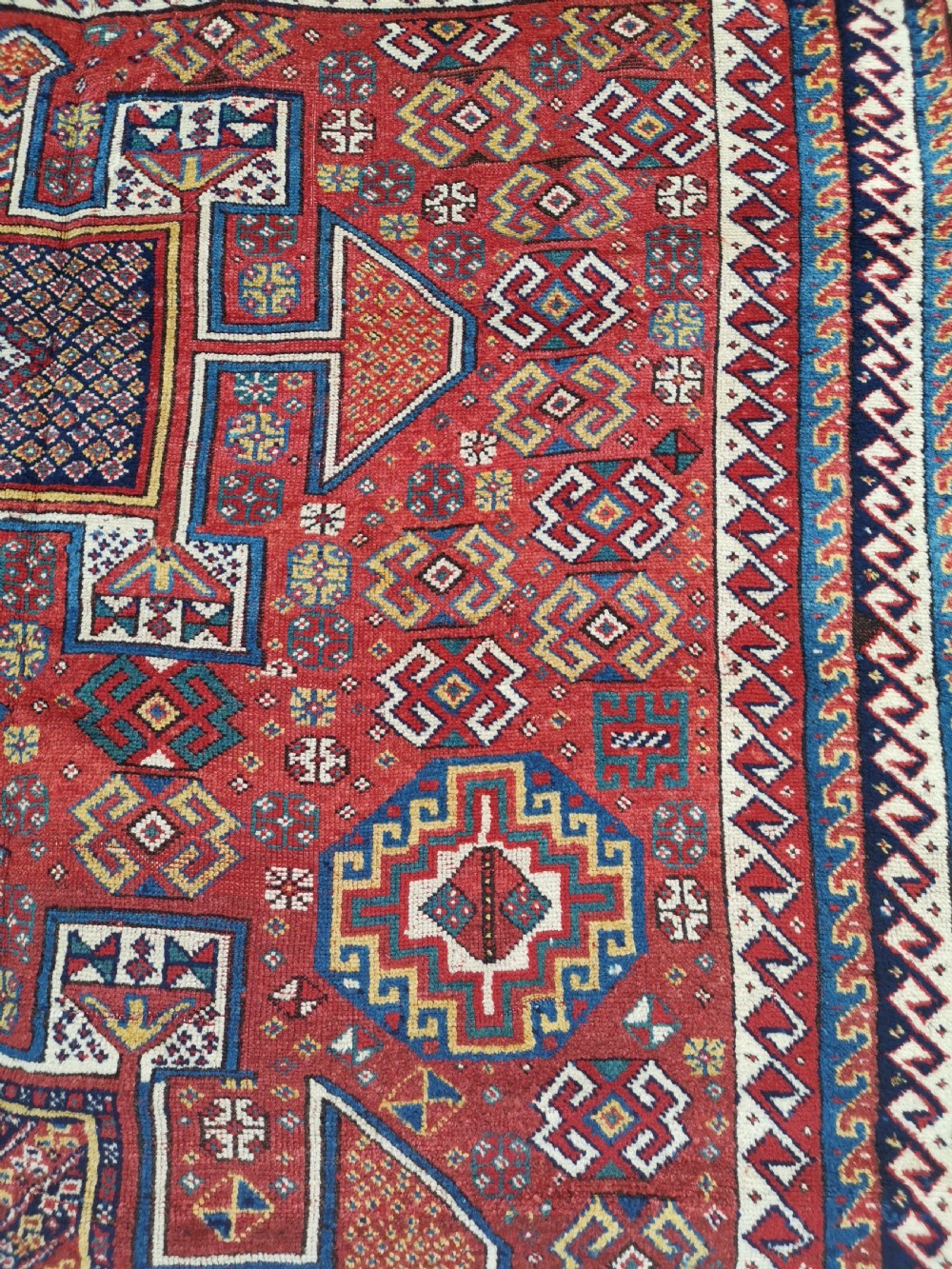 A handwoven rectangular Quchan Kurdish wool rug featuring geometric stepped medallions and repeating motifs in shades of terracotta red, navy blue, and cream. View V
