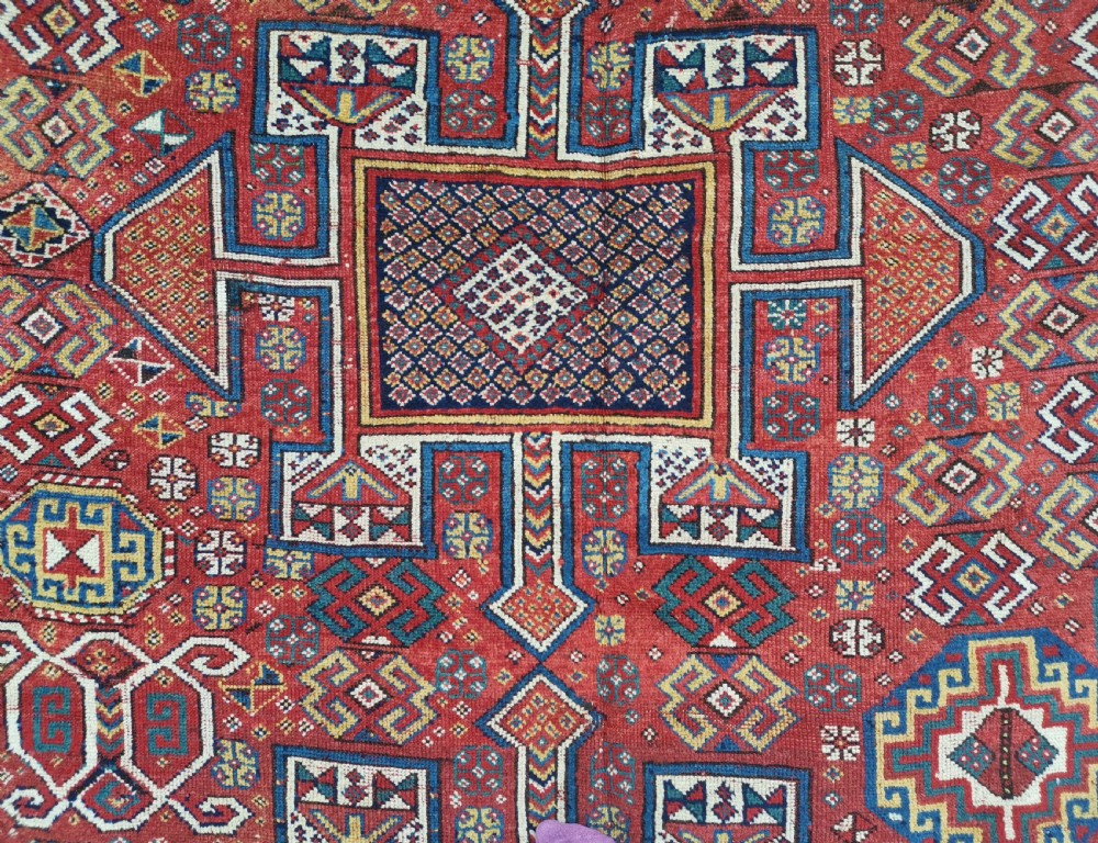 A handwoven rectangular Quchan Kurdish wool rug featuring geometric stepped medallions and repeating motifs in shades of terracotta red, navy blue, and cream. View X