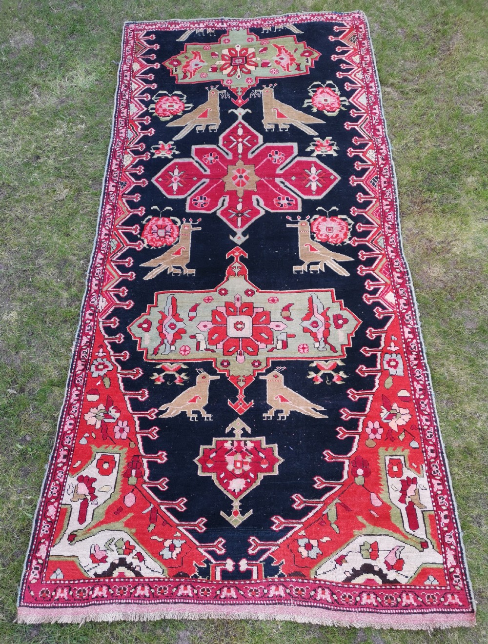 A detailed close-up view of an antique dark navy hand-knotted wool rug featuring stylized bird figures, red geometric star medallions, and floral motifs. View II