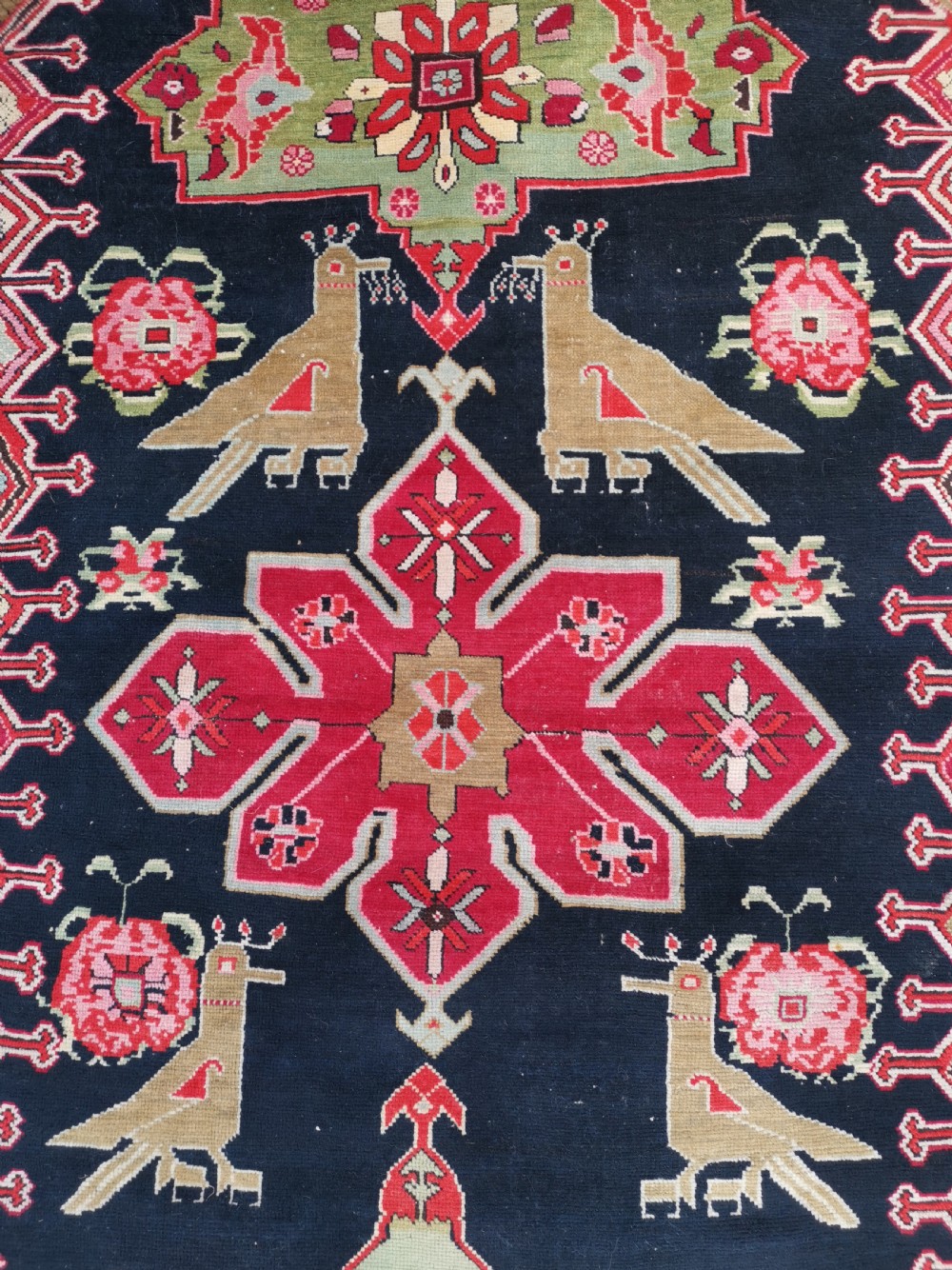 A detailed close-up view of an antique dark navy hand-knotted wool rug featuring stylized bird figures, red geometric star medallions, and floral motifs. View III