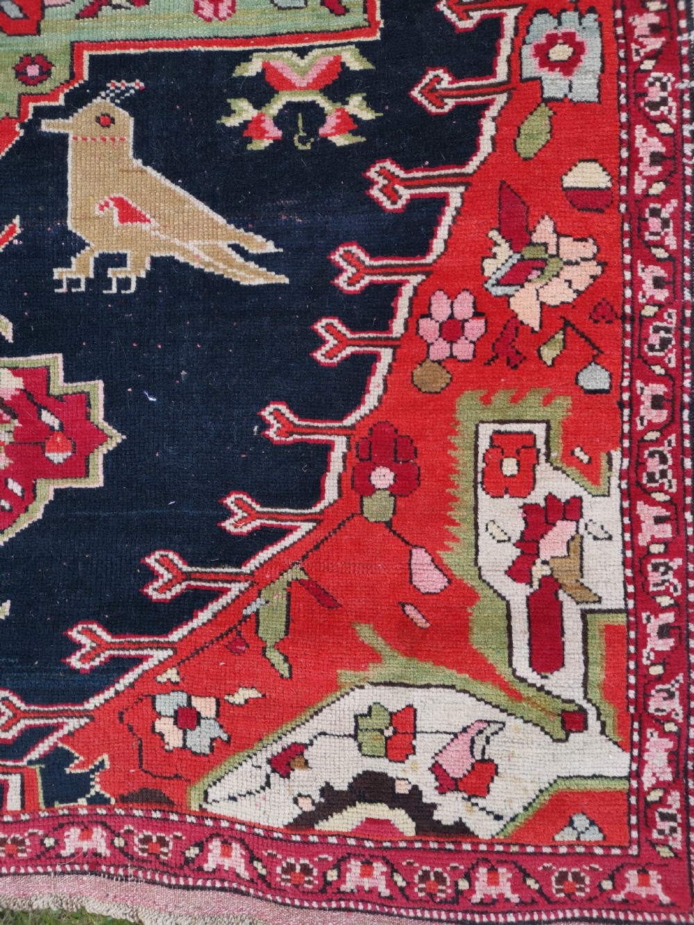 A detailed close-up view of an antique dark navy hand-knotted wool rug featuring stylized bird figures, red geometric star medallions, and floral motifs. View V