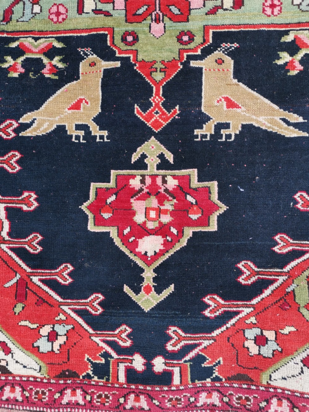 A detailed close-up view of an antique dark navy hand-knotted wool rug featuring stylized bird figures, red geometric star medallions, and floral motifs. View VI