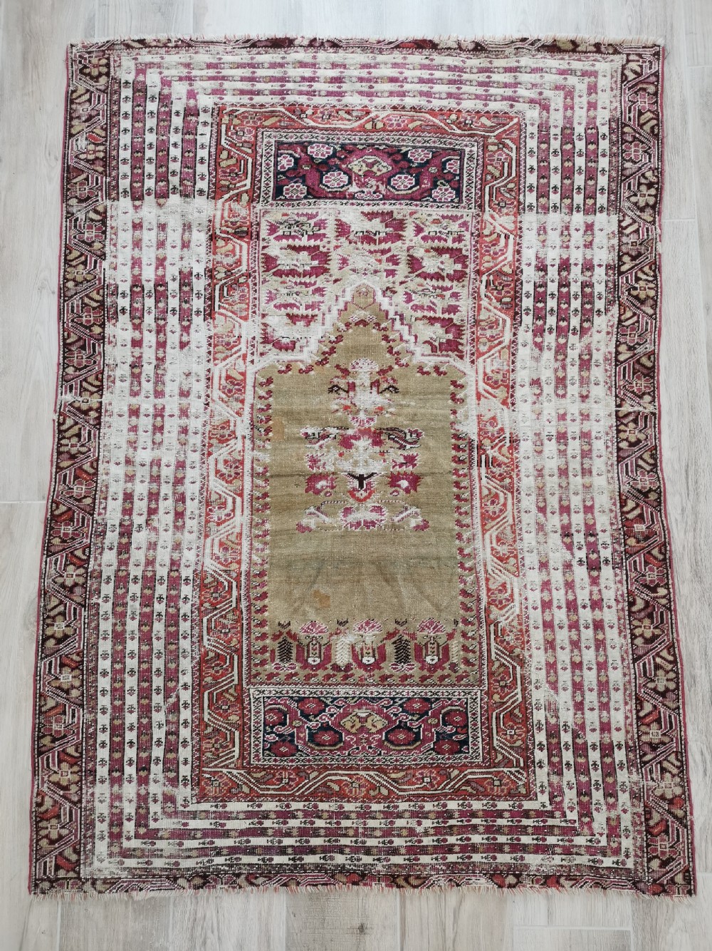 An antique Anatolian Ghiordes prayer rug featuring a central beige arched mihrab panel framed by multiple geometric borders in crimson red and cream wool.