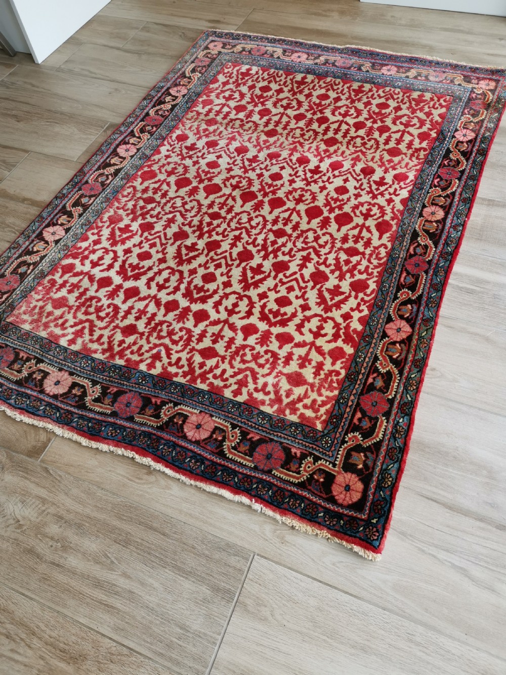 A hand-knotted rectangular antique Sarouk wool rug featuring a crimson red repeating floral pattern on a cream ground framed by a dark floral border. View VI