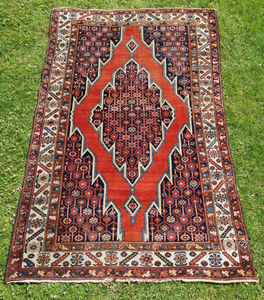 An antique Persian Mazlaghan wool rug featuring a central stepped navy and cream diamond medallion on a terracotta red field with geometric borders.