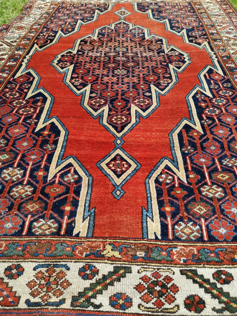 An antique Persian Mazlaghan wool rug featuring a central stepped navy and cream diamond medallion on a terracotta red field with geometric borders. View III