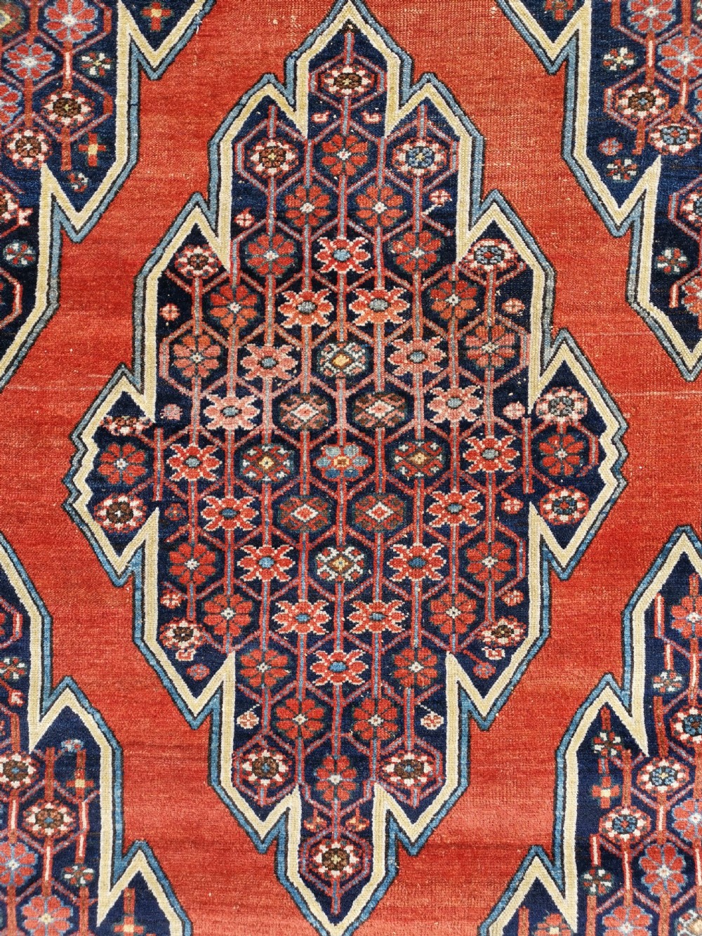 An antique Persian Mazlaghan wool rug featuring a central stepped navy and cream diamond medallion on a terracotta red field with geometric borders. View IV