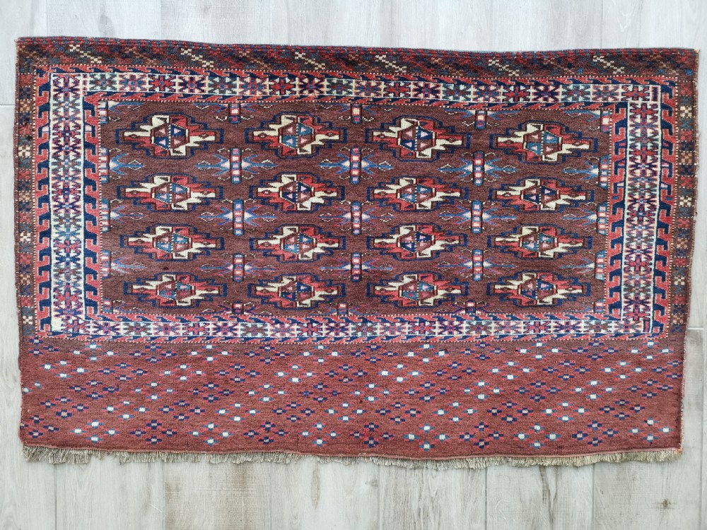 A rectangular hand-knotted wool Yomut chuval featuring rows of geometric guls on a reddish brown field above a patterned lower elem panel.