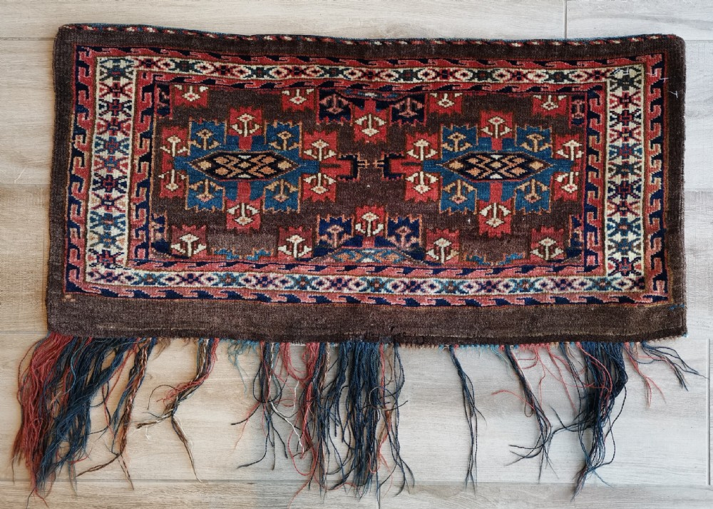 A rectangular hand-knotted wool Yomut torba featuring a dark brown background, geometric red and blue guls, a patterned border, and loose wool fringe along the bottom edge.